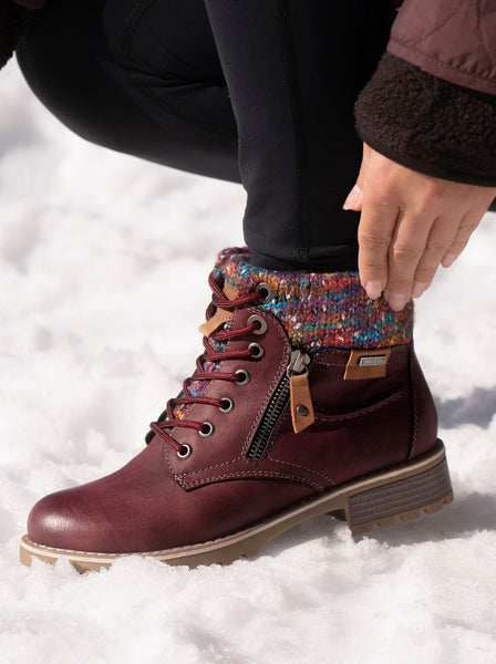A close-up of a person's hand adjusting a colorful knit ankle cuff on a burgundy leather lace-up boot, set against a snowy outdoor background.