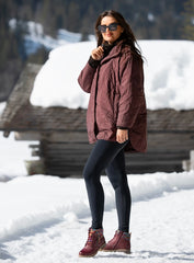 A woman wearing a burgundy quilted winter jacket, black leggings, and burgundy lace-up Lunar waterproof boots stands in a snowy outdoor setting with a rustic wooden cabin in the background.