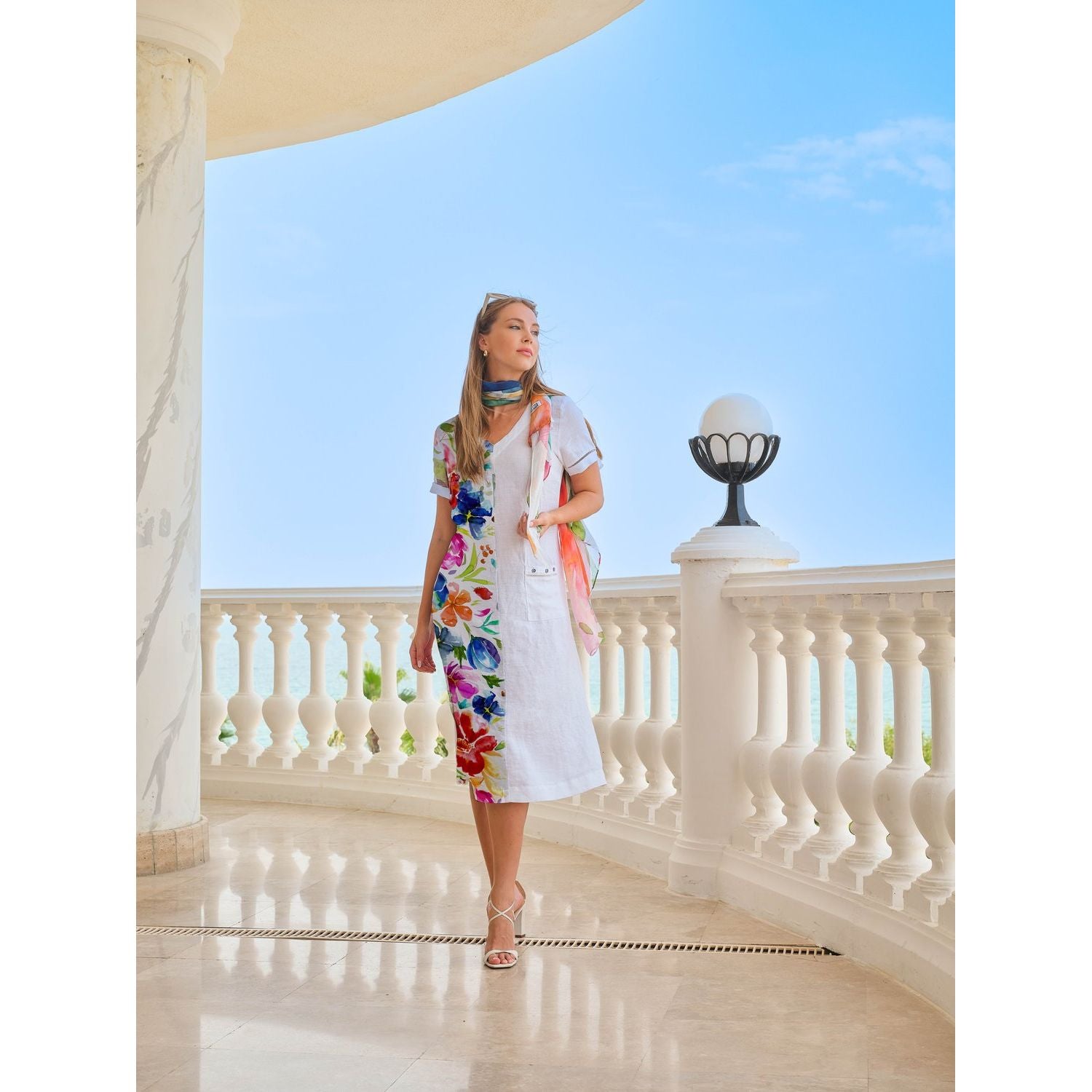 Woman in a floral dress standing on a balcony with a clear blue sky.