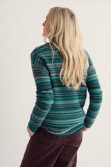 A woman with long blonde hair is shown from the back wearing a green and teal striped knit sweater and dark brown corduroy pants against a plain off-white background.