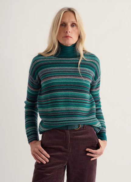 A woman wearing a green and teal Seasalt striped turtleneck sweater with dark brown corduroy pants, standing against a plain off-white studio background.