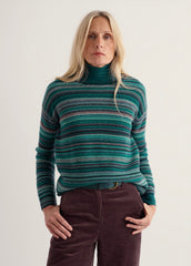 A woman wearing a green and teal Seasalt striped turtleneck sweater with dark brown corduroy pants, standing against a plain off-white studio background.