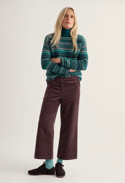 A woman stands against a plain light background wearing a green and teal striped turtleneck sweater, dark brown wide-leg corduroy pants, light blue socks, and black shoes.