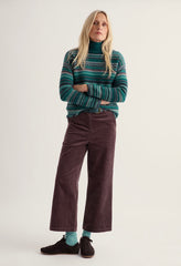A woman stands against a plain light background wearing a green and teal striped turtleneck sweater, dark brown wide-leg corduroy pants, light blue socks, and black shoes.