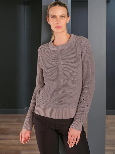 A woman modeling a taupe metalized knit crewneck sweater with decorative sleeve cutouts, paired with black pants, set against a dark gray modern interior background.