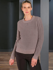 A woman modeling a taupe metalized knit crewneck sweater with decorative sleeve cutouts, paired with black pants, set against a dark gray modern interior background.