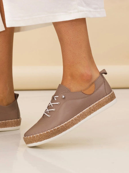 A person wearing Lunar taupe-brown slip-on espadrille-style shoes with white laces, a cork-textured midsole, and white soles, shown against a warm beige studio background.