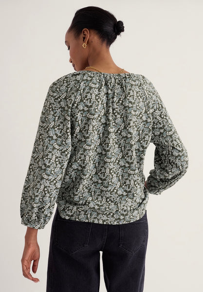 A woman shown from the back wearing a long-sleeve blouse with a dense abstract floral or paisley print in muted green, gray, and off-white tones, paired with dark pants, against a plain light background.
