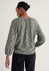 A woman shown from the back wearing a long-sleeve blouse with a dense abstract floral or paisley print in muted green, gray, and off-white tones, paired with dark pants, against a plain light background.