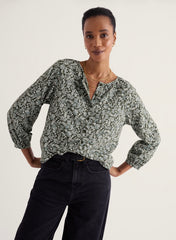 A woman modeling a long-sleeve blouse with a dense abstract print in muted green, gray, and white, paired with dark jeans against a plain light background.