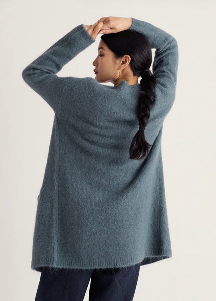 A woman wearing a Seasalt, oversized long-sleeve sweater in a muted slate blue-gray color, shown from the back against a plain light background.