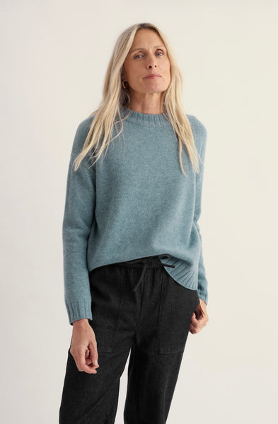 A woman modeling a light blue knit sweater with black pants against a plain off-white studio background.