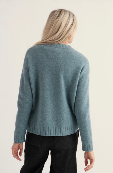 A woman viewed from the back wearing a Seasalt blue knit sweater and black pants against a plain light background.