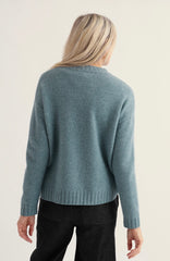 A woman viewed from the back wearing a Seasalt blue knit sweater and black pants against a plain light background.