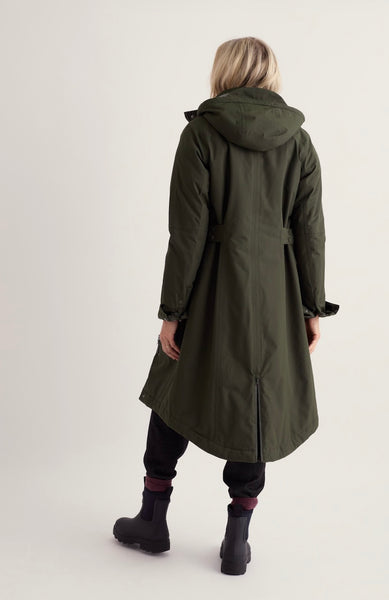 A woman viewed from behind wearing a long olive green hooded parka coat, black pants, and black boots against a plain light gray studio background.