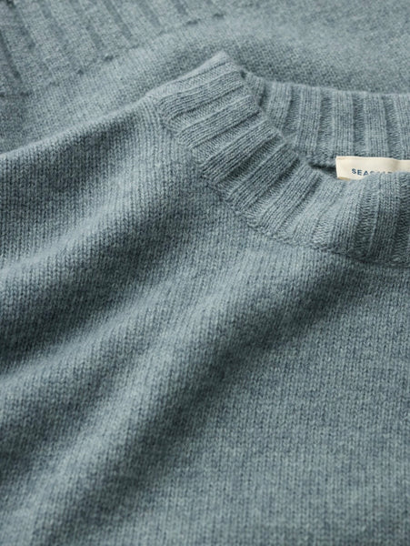 Close-up of a Seasalt knit sweater in a muted dusty blue-gray color, showing the texture of the fabric and a small white brand tag near the collar.