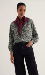 A woman modeling a Seasalt long-sleeve button-up blouse with a green and white floral pattern, styled with a deep purple neck scarf and dark wide-leg jeans against a plain light background.