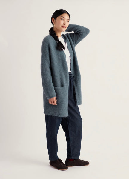 A woman modeling a long, Seasalt open-front cardigan in a muted blue-gray color, styled with a white top, dark blue jeans, and dark brown slip-on shoes against a plain light background.