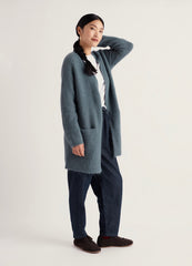 A woman modeling a long, Seasalt open-front cardigan in a muted blue-gray color, styled with a white top, dark blue jeans, and dark brown slip-on shoes against a plain light background.