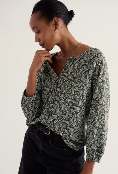 A woman wearing a long-sleeve V-neck blouse with a green and white abstract floral pattern, paired with black jeans, photographed against a plain light background.