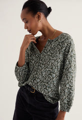 A woman wearing a long-sleeve V-neck blouse with a green and white abstract floral pattern, paired with black jeans, photographed against a plain light background.