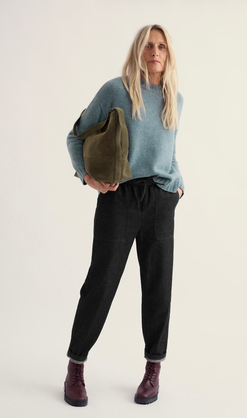 A woman stands against a plain light background wearing a light blue knit Seasalt sweater, dark charcoal pants, burgundy boots, and holding a large olive green tote bag.