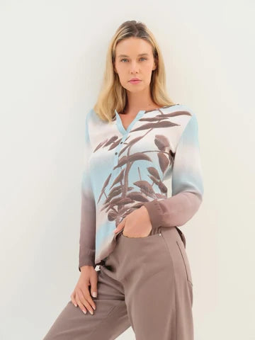 A woman modeling a long-sleeve V-neck top with a light blue, gray, and brown botanical leaf print, paired with taupe trousers, against a plain off-white background.