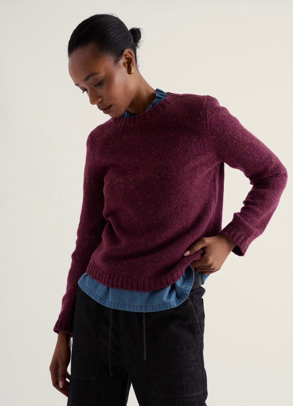 A woman wearing a heathered burgundy knit sweater over a blue collared shirt and dark charcoal pants, photographed against a plain light beige studio background.