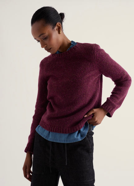 A woman wearing a heathered burgundy knit sweater over a blue collared shirt and dark charcoal pants, photographed against a plain light beige studio background.