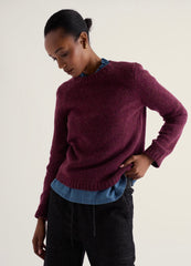 A woman wearing a heathered burgundy knit sweater over a blue collared shirt and dark charcoal pants, photographed against a plain light beige studio background.