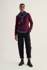 A model stands against a plain light beige studio background wearing a burgundy knit  seasalt sweater over a blue shirt, a blue neck scarf, dark charcoal pants, and black boots.