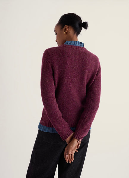A woman shown from the back wearing a textured knit sweater in a deep plum purple, layered over a blue ruffled shirt and paired with black pants, against a plain off-white background.