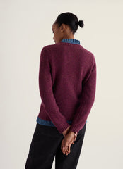A woman shown from the back wearing a textured knit sweater in a deep plum purple, layered over a blue ruffled shirt and paired with black pants, against a plain off-white background.